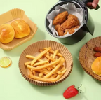 A tabletop food scene on a pale mint background showing an air fryer filled with golden fried chicken wings, paper basket-style plates with fries and bread rolls, and a strawberry on the surface.