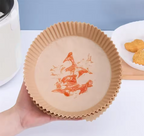A hand holds a round brown Air Fryer Paper Tray with a printed red fish design in the center. The background shows a kitchen countertop with a white appliance on the left and a plate of food on the right.