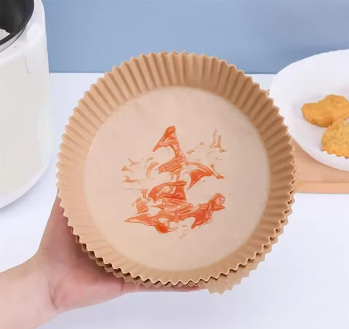 A hand holds a round brown Air Fryer Paper Tray with a printed red fish design in the center. The background shows a kitchen countertop with a white appliance on the left and a plate of food on the right.