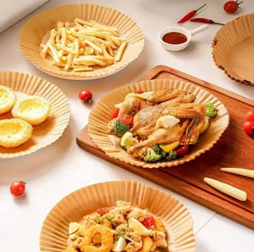 A styled food presentation featuring a whole roasted chicken with broccoli and peppers served on a brown paper plate, alongside french fries, mini tarts, and other dishes on matching Air Fryer Paper Tray and a wooden board.